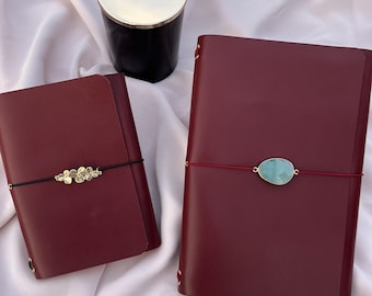 May include: Two burgundy leather journals with decorative accents. One journal features a gold floral embellishment on a black band, the other has a light blue gemstone on a red band. A black candle with a gold rim sits in the background. The text "Burgundy Elegance" is at the bottom.