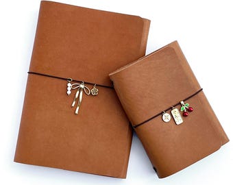 May include: Two brown leather journals of different sizes, secured with elastic bands. The larger journal has a gold bow charm, while the smaller one features charms of a cherry, a compass, and a tag. The journals are set against a white background.