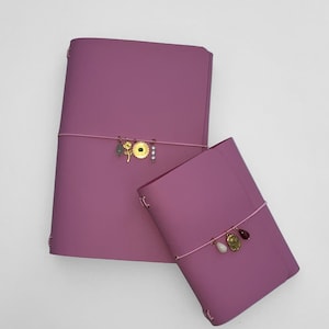 Op de afbeelding: Twee mauve leren journals van verschillende formaten, vastgezet met roze elastiekjes. Elk journal heeft decoratieve gouden bedels en kralen. Het grotere journal heeft een gouden bloem bedel en de kleinere een gouden munt bedel.