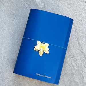 Könnte beinhalten: Ein leuchtend blaues Notizbuch mit dem in Gold geprägten Schriftzug Made of Moments auf dem Vorderdeckel. Ein dünnes blaues Gummiband umschließt die Mitte und hält einen blassgelben, durchscheinenden, blumenförmigen Anhänger.