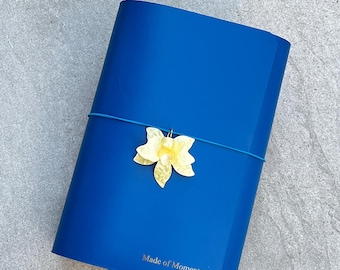 May include: A vibrant blue notebook featuring the text Made of Moments embossed in gold on the front cover. A thin blue elastic band wraps around the center, holding a pale yellow, translucent, flower-shaped charm in place.