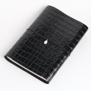 May include: A black notebook with a glossy crocodile-embossed texture. The cover features a small, centered gold-toned charm with a white teardrop-shaped pearl. The notebook is closed, revealing white pages along the bottom edge of the binding.