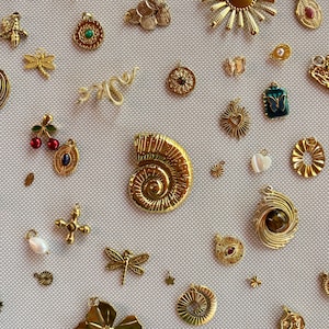 Può includere: Una collezione di ciondoli in metallo color oro di varie forme, tra cui un sole, un grande fossile di ammonite, libellule, fiori, una ciliegia, un serpente e piccoli pendenti geometrici con pietre incastonate e dettagli in perla bianca.