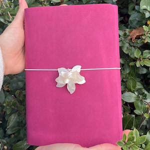 May include: A magenta journal with a white floral decoration and a white elastic closure. The journal is held against a backdrop of green leaves. The floral decoration is centrally placed on the journal.