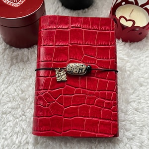 Op de afbeelding: Een rood leren notitieboek met een krokodillenleertextuur. Het notitieboek is vastgezet met een zwarte elastische band en een decoratieve bedel. Een kleine, rode, hartvormige doos en een kandelaar staan op de achtergrond.