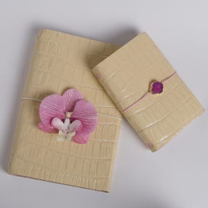May include: Two rectangular journals with a cream-colored, embossed crocodile-skin texture. The larger journal features a pink orchid flower attached to a white elastic band. The smaller journal has a pink elastic band with a gold-rimmed magenta stone.