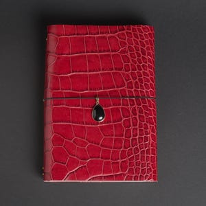 May include: A rectangular journal with a vibrant red cover featuring a textured crocodile skin pattern. A thin black elastic band wraps horizontally around the centre of the cover, securing a small, smooth, black teardrop-shaped pendant charm.