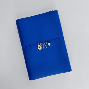 May include: A vibrant royal blue notebook secured with a matching elastic band. Three small gold-toned charms are attached to the elastic: a circular medallion with a central blue stone, a delicate bow shape, and a single iridescent white pearl bead.