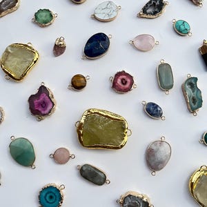 Puede incluir: Una colección de colgantes y conectores de piedras preciosas variadas con diversas formas, tamaños y colores, incluyendo morado, verde, azul, rosa y blanco. Cada pieza está rodeada por un borde metálico dorado e incluye pequeñas anillas.