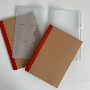 Op de afbeelding: Twee notitieboeken met bruine kaften en rode stoffen ruggen liggen naast een documentenmap van doorschijnend plastic. De map is voorzien van drie interne vakken voor kaarten en een witte ritssluiting aan de rechterkant.