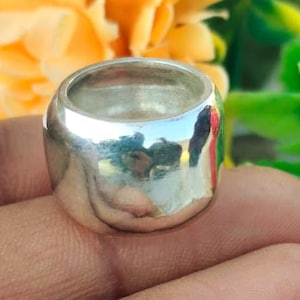May include: A chunky, polished silver ring with a rounded shape. The ring reflects the surrounding environment, creating a mirrored effect. The ring is held in a hand, with a blurred background of yellow and green foliage.