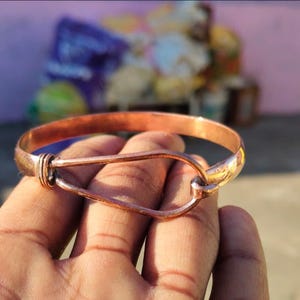 Puede incluir: Una pulsera de color cobre con un cierre único de gancho y ojo. La pulsera es una banda simple con un diseño de gancho curvo y alargado. El metal tiene un tono cálido marrón rojizo, y la pulsera parece hecha a mano. La pulsera se sostiene en una mano.
