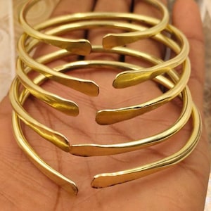 May include: A stack of six gold-colored, open-ended bangles. Each bangle has a smooth, rounded band that tapers slightly at the open ends. The bangles are stacked on top of each other, creating a layered effect. The metal has a polished, reflective surface.