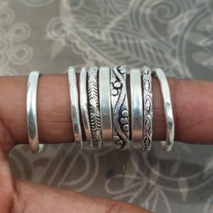 Può includere: Un set di otto anelli color argento con vari design, tra cui fasce lisce, texture martellate e motivi complessi con linee incise, punti e disegni a forma di foglia, tutti mostrati insieme su un dito per dare un'idea delle dimensioni.