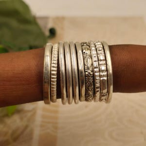 May include: A collection of silver rings of varying widths and designs. Some rings are smooth, while others feature textured patterns, including ridges and floral motifs. The rings are stacked together, creating a layered effect.