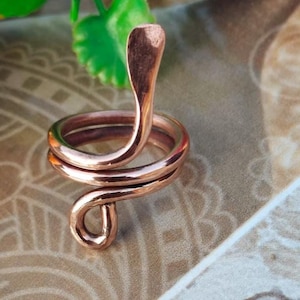 May include: A copper-coloured ring shaped like a cobra snake. The snake's head is raised, and the body coils around the finger. The ring is made of a shiny, metallic material, likely a piece of jewellery.