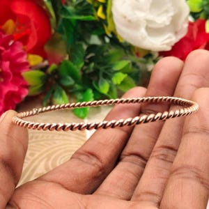 Op de afbeelding: Een roségouden, gedraaide armband. De armband heeft een doorlopend touwachtig ontwerp. De achtergrond toont kleurrijke bloemen.