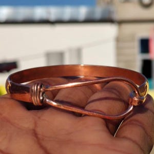 Puede incluir: Un brazalete de color cobre con cierre de gancho. El brazalete está hecho de una sola banda de cobre con un diseño simple y elegante. El cierre de gancho es una característica única que agrega un toque de estilo a la pieza.