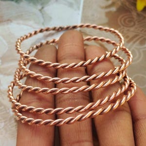 May include: Several copper-colored, twisted-wire bangles are stacked on top of each other. The bangles have a spiral design and are made of a shiny, metallic material. The bracelets are displayed on a person's fingers.