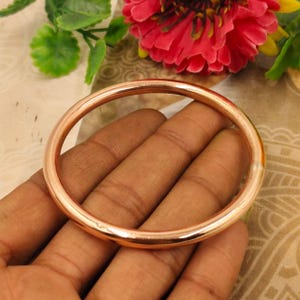 May include: A copper-colored bangle bracelet. The round bracelet is held in a hand, with a blurred background of flowers and foliage. The bracelet has a smooth, polished surface and a simple, minimalist design.