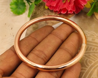 May include: A copper-colored bangle bracelet. The round bracelet is held in a hand, with a blurred background of flowers and foliage. The bracelet has a smooth, polished surface and a simple, minimalist design.
