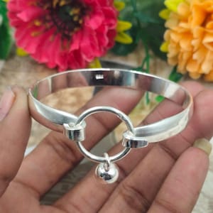 May include: A silver-colored bangle bracelet with a circular design and a small dangling ball. The bracelet has a smooth, polished finish and a simple, elegant design. The bracelet is shown against a background of colorful flowers.