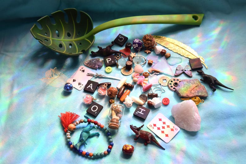 May include: A collection of small decorative items, including a green leaf-shaped serving spoon, playing cards, dice, and miniature figurines. The items are arranged on a light blue surface, creating a whimsical and eclectic display.