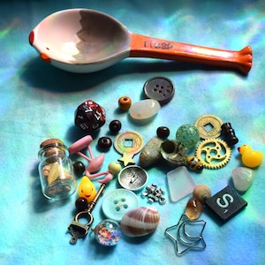 May include: A collection of small objects including a measuring spoon, dice, buttons, beads, a miniature glass jar, a rubber duck, and various charms. The spoon is white with an orange handle and the text "1 TSP".