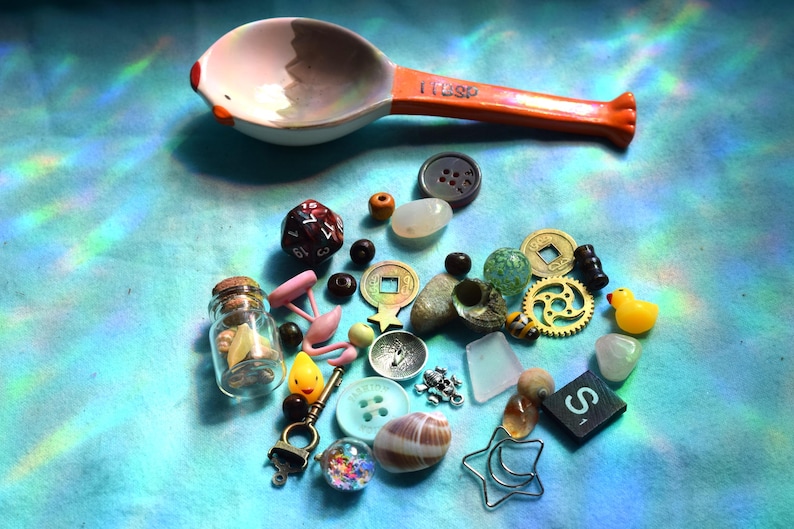 May include: A collection of small objects including a measuring spoon, dice, buttons, beads, a miniature glass jar, a rubber duck, and various charms. The spoon is white with an orange handle and the text "1 TSP".