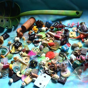 May include: A collection of small decorative items, including dice, buttons, miniature flamingos, corks, and shells, scattered on a blue surface. A green leaf-shaped spoon is at the top. Various colors and textures are visible.