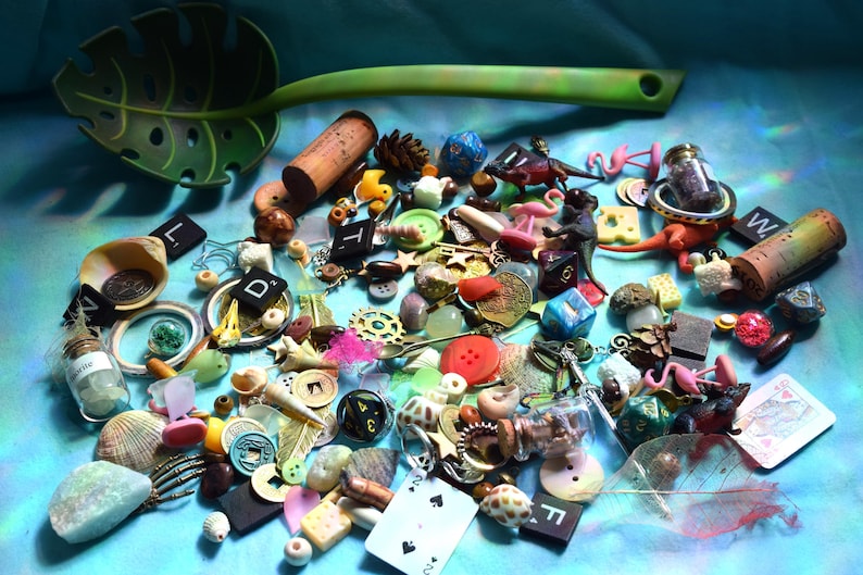 May include: A collection of small decorative items, including dice, buttons, miniature flamingos, corks, and shells, scattered on a blue surface. A green leaf-shaped spoon is at the top. Various colors and textures are visible.