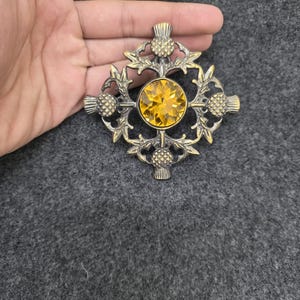May include: A vintage brooch with a thistle design. The brooch features a large, round, yellow gemstone at the center, surrounded by an ornate, gold-toned metal frame. The frame is shaped like a cross with thistle motifs.