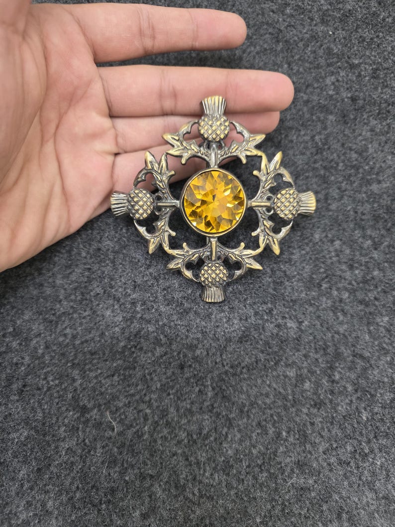 May include: A vintage brooch with a thistle design. The brooch features a large, round, yellow gemstone at the center, surrounded by an ornate, gold-toned metal frame. The frame is shaped like a cross with thistle motifs.