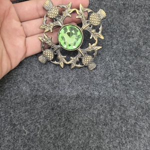 May include: A vintage-style brooch with a central, round, light green gemstone. The brooch features a detailed design of thistles and leaves in a bronze-colored metal. The brooch is square-shaped and has a pin on the back.