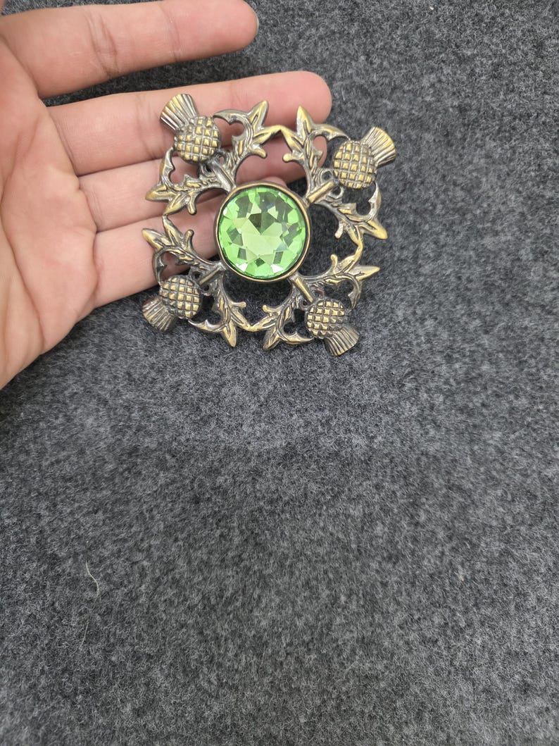 May include: A vintage-style brooch with a central, round, light green gemstone. The brooch features a detailed design of thistles and leaves in a bronze-colored metal. The brooch is square-shaped and has a pin on the back.