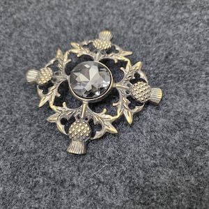 May include: A vintage-style brooch with a central gray gemstone, surrounded by a detailed, gold-toned metal design. The design incorporates thistle motifs and intricate leaf patterns, creating a classic and elegant accessory.