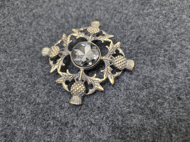 May include: A vintage-style brooch with a central gray gemstone, surrounded by a detailed, gold-toned metal design. The design incorporates thistle motifs and intricate leaf patterns, creating a classic and elegant accessory.
