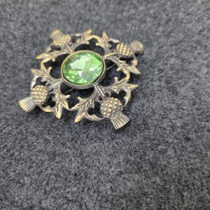 May include: Vintage brooch with a thistle design. The brooch features a central oval green gemstone surrounded by a detailed gold-toned thistle motif. The brooch is a square shape with four thistles at each corner.