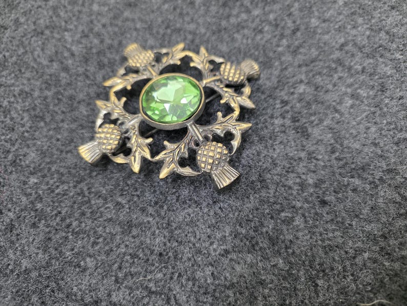 May include: Vintage brooch with a thistle design. The brooch features a central oval green gemstone surrounded by a detailed gold-toned thistle motif. The brooch is a square shape with four thistles at each corner.