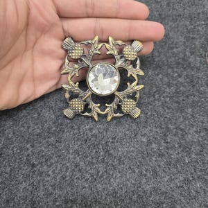 May include: A square, antique-style brooch with a central, clear, round gemstone. The brooch features a thistle design in a gold-colored metal. The thistle design is on each corner of the square.