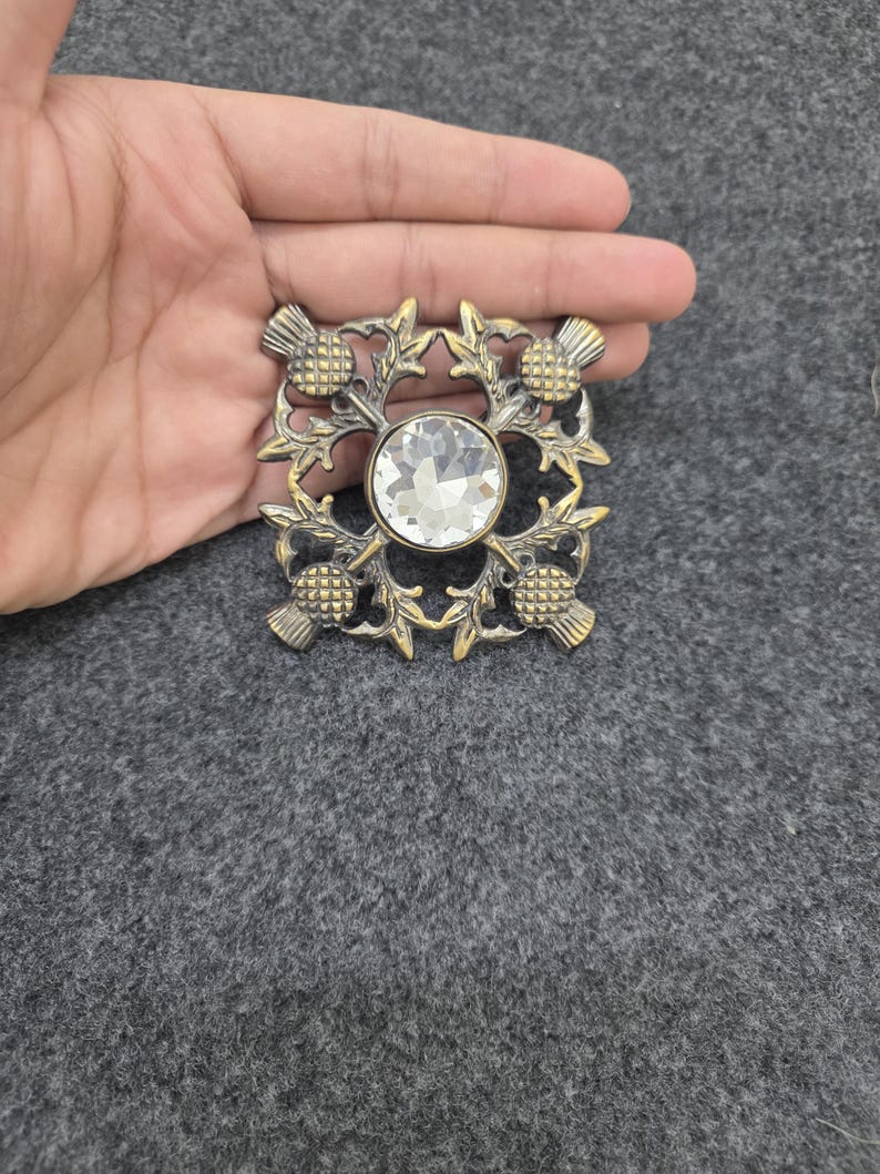 May include: A square, antique-style brooch with a central, clear, round gemstone. The brooch features a thistle design in a gold-colored metal. The thistle design is on each corner of the square.