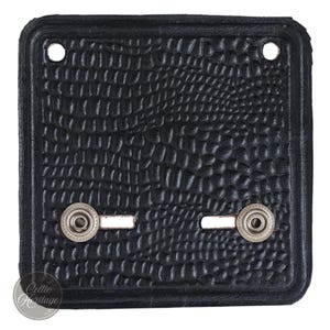 May include: A square black leather patch featuring a textured crocodile skin pattern. The patch includes two circular holes at the top corners and two silver-toned metal snap fasteners positioned horizontally in the lower section of the piece.