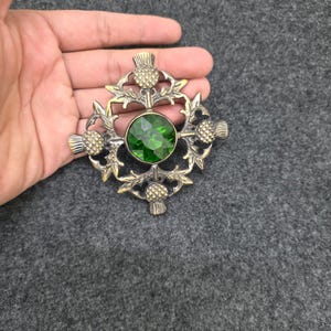 May include: A vintage-style brooch with a central, round, emerald-green gemstone. The brooch features a detailed thistle design in a gold-toned metal, with intricate leaves and thistle heads. The brooch is held in a hand.