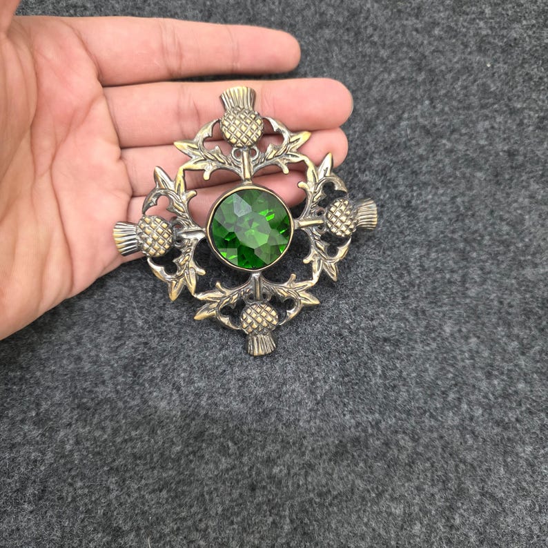 May include: A vintage-style brooch with a central, round, emerald-green gemstone. The brooch features a detailed thistle design in a gold-toned metal, with intricate leaves and thistle heads. The brooch is held in a hand.