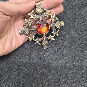 May include: A bronze-toned brooch with a central, round, orange-red gemstone. The brooch features a thistle design with intricate detailing. The design includes four thistles around the gemstone and decorative leaves.