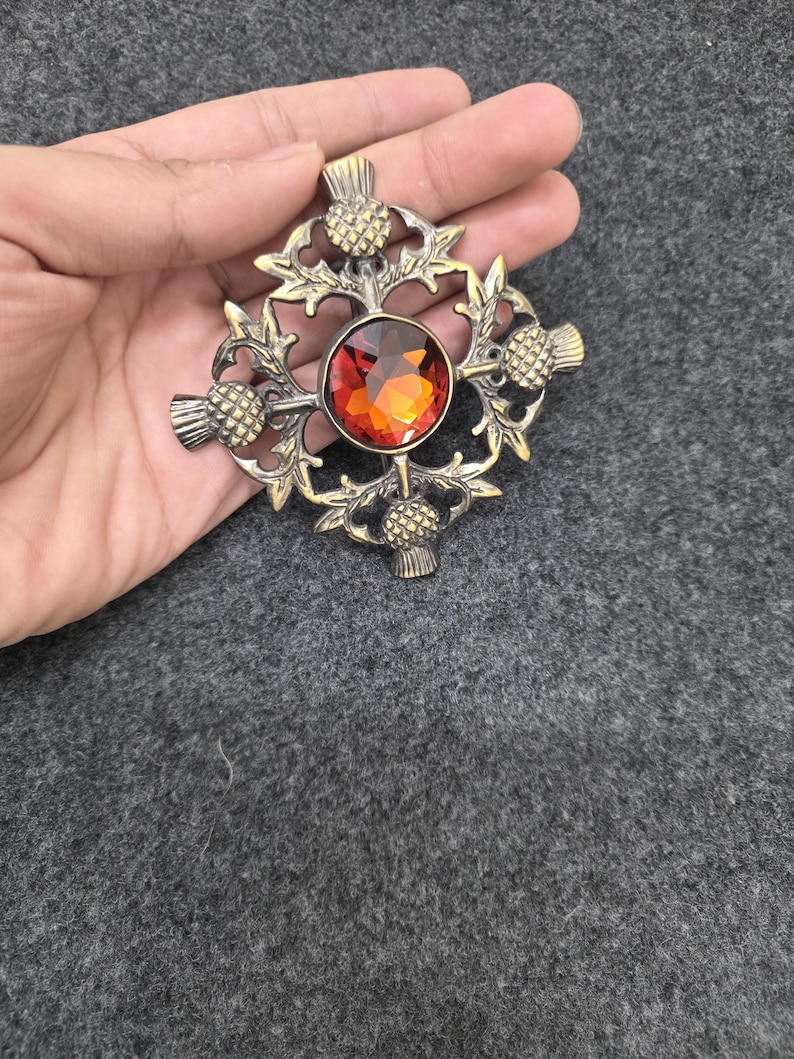May include: A bronze-toned brooch with a central, round, orange-red gemstone. The brooch features a thistle design with intricate detailing. The design includes four thistles around the gemstone and decorative leaves.