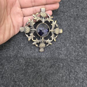 May include: A bronze-toned brooch with a central dark purple gemstone, surrounded by a decorative thistle and leaf design. The brooch has a cross-like shape with thistle accents at each end.