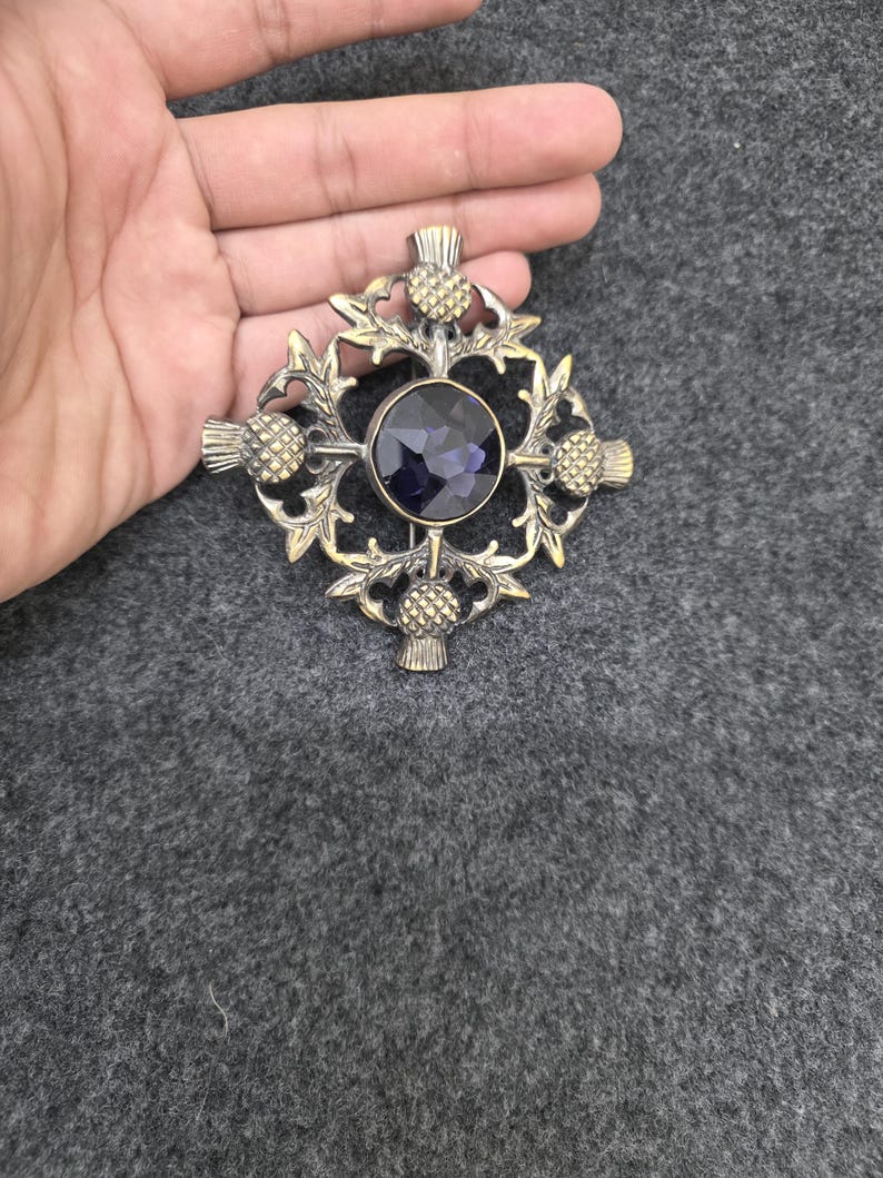 May include: A bronze-toned brooch with a central dark purple gemstone, surrounded by a decorative thistle and leaf design. The brooch has a cross-like shape with thistle accents at each end.
