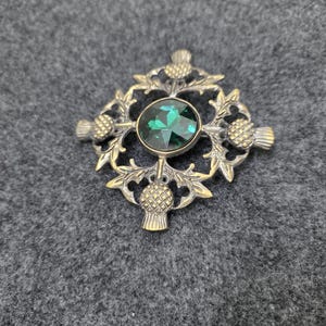 May include: A silver-toned brooch with a central emerald-green gemstone. The brooch features a detailed design of thistles and leaves, with a round gemstone in the center. The brooch is set against a gray background.