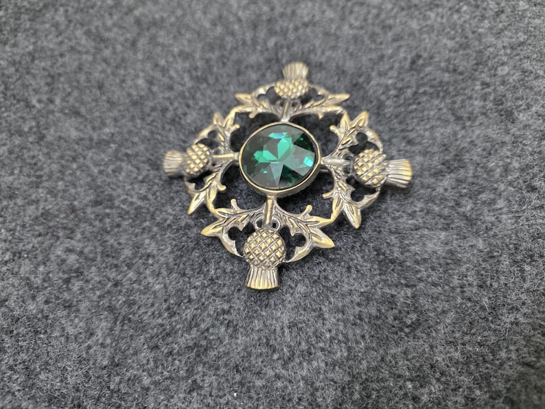 May include: A silver-toned brooch with a central emerald-green gemstone. The brooch features a detailed design of thistles and leaves, with a round gemstone in the center. The brooch is set against a gray background.